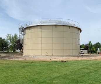 Large bolted steel water storage tank with exterior stair access, installed on a landscaped site for municipal or industrial use