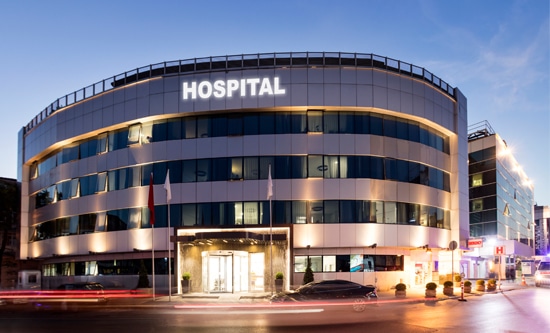Modern hospital building exterior with curved architecture, glass windows, and illuminated hospital sign at dusk
