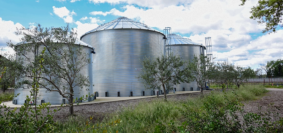 Corrugated steel water storage tanks surrounded by native landscaping and olive trees, designed for sustainable irrigation and rainwater harvesting